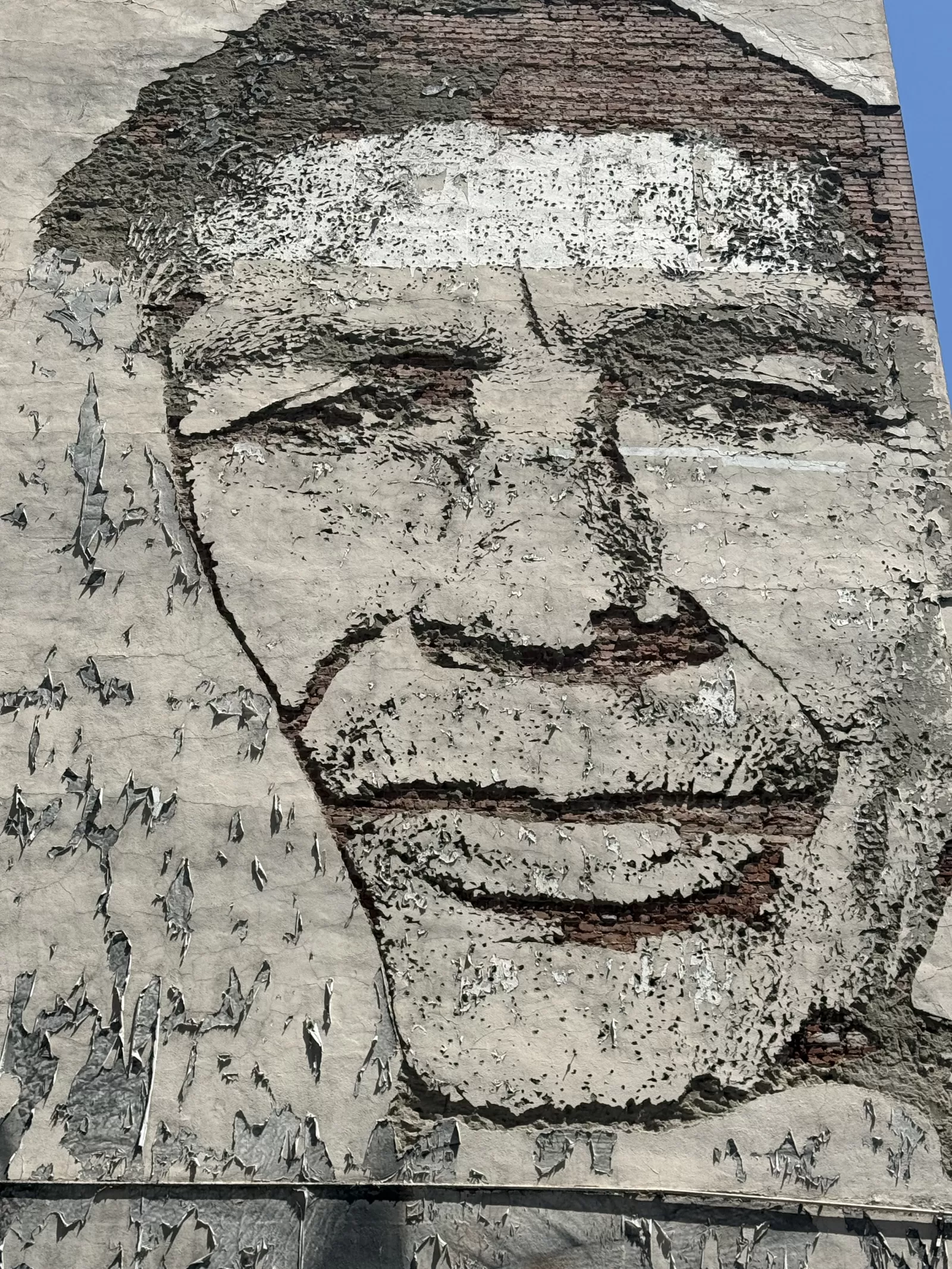 A large monochrome portrait is painted across an exterior wall, with facial features formed by dark outlines and exposed reddish brick. Extensive peeling, cracking, and weathering reveal layers of plaster and masonry beneath the image.