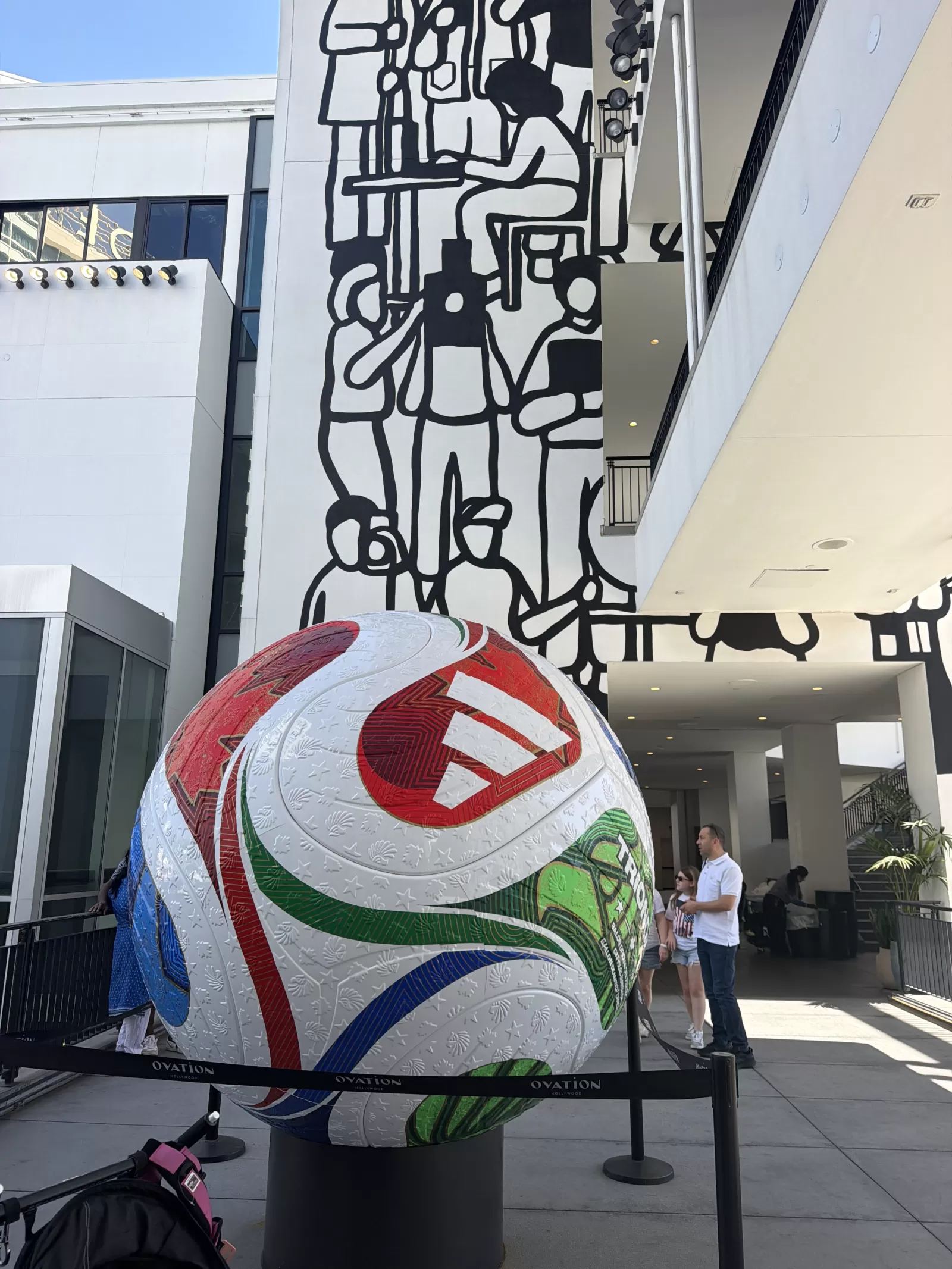 A large black-and-white mural covers the exterior wall of a modern building, featuring interconnected abstract, human-like outlined forms and geometric shapes. A large colorful patterned sphere is displayed in front of the mural, with several people visible nearby.
