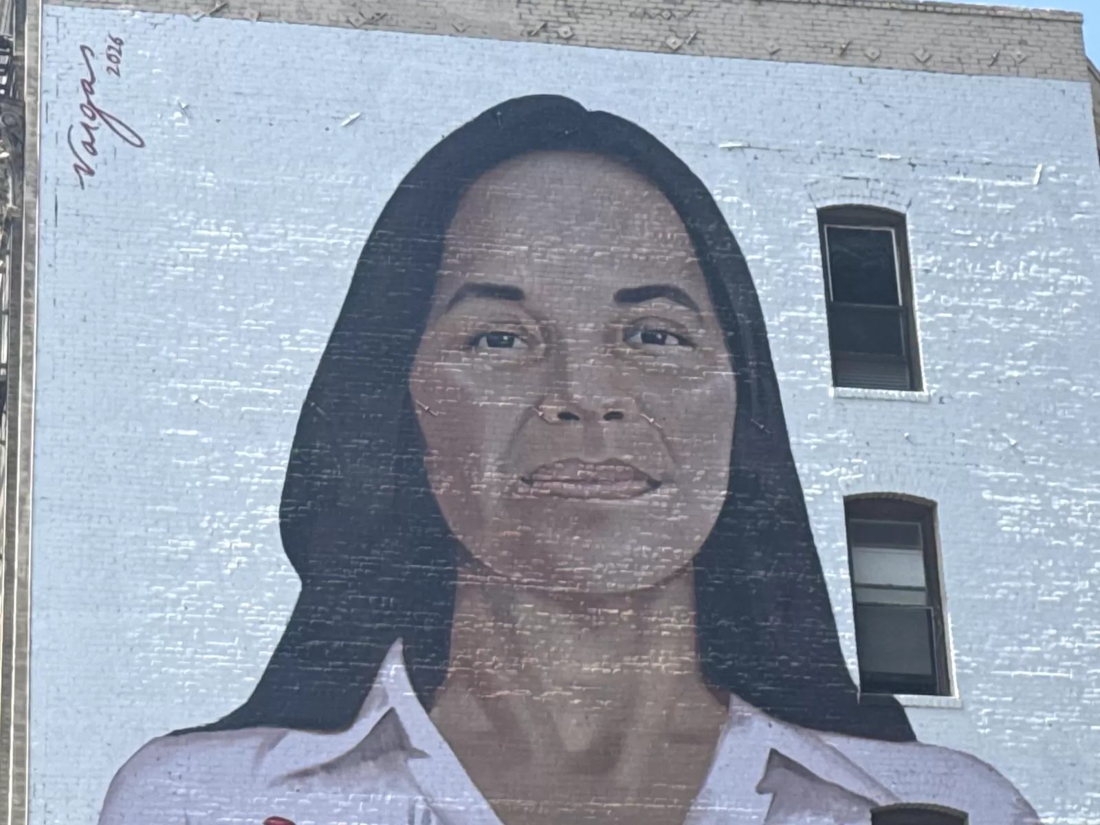 A large painted portrait of a woman with long dark hair and a light-colored collared shirt covers the blue-painted brick wall of a building. A small handwritten signature and the date “2026” are visible near the upper left, with windows integrated into the wall on the right.