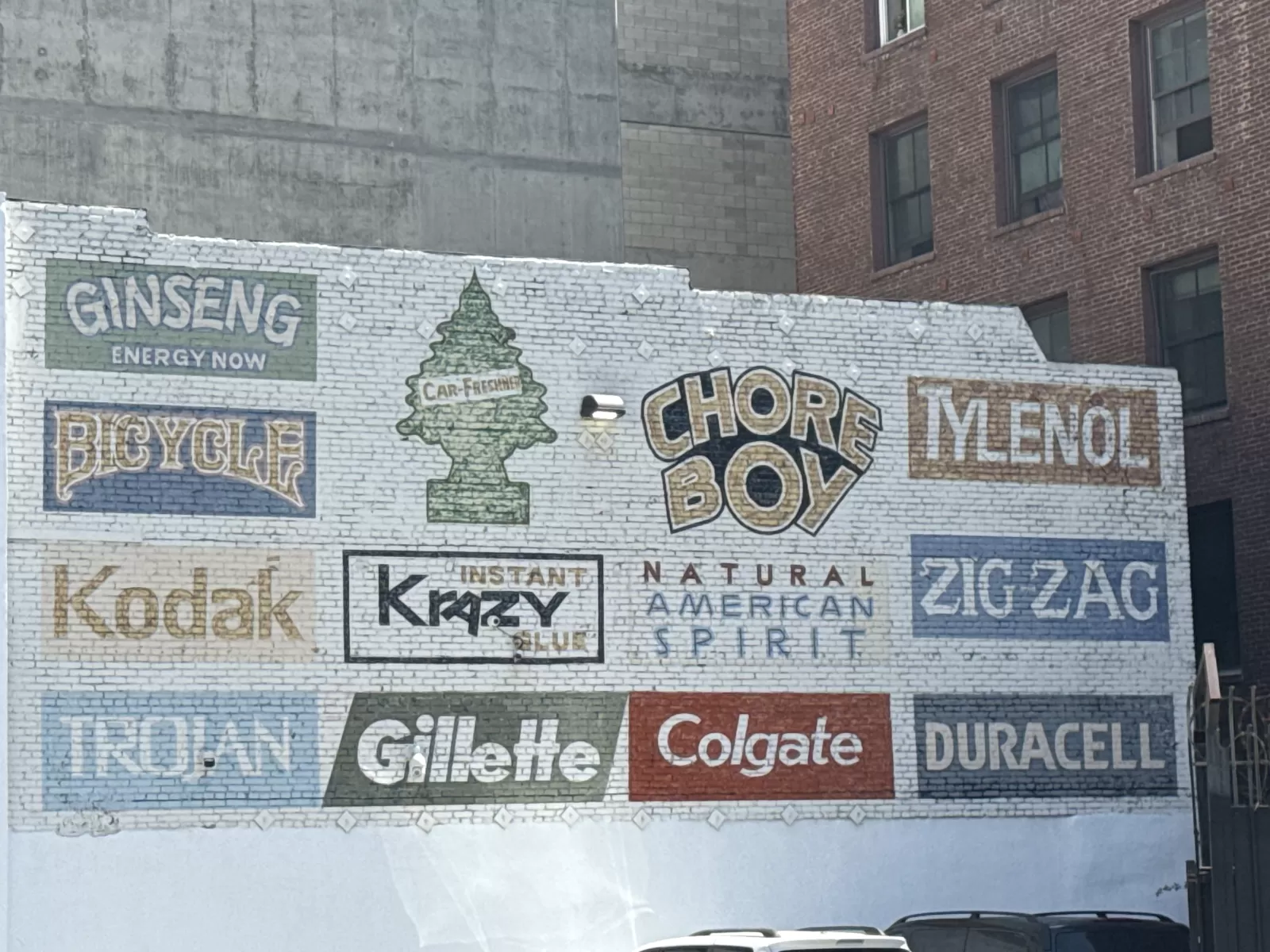 A painted brick wall displays vintage-style product lettering and logos, including “Ginseng Energy Now,” “Bicycle,” “Chore Boy,” “MENOL,” “Kodak,” “Krazy,” “Gillette,” “Colgate,” “Duracell,” and “Zig Zag.” The mural is beside a brick building and includes a central evergreen-shaped graphic labeled “Car-Freshner.”