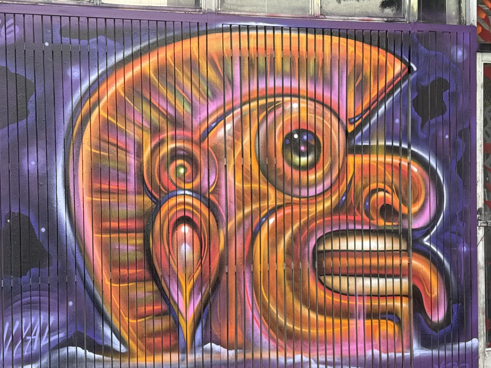 A large abstract mural covers a barred exterior wall in vivid orange, pink, purple, yellow, black, and white forms. The composition includes overlapping curves, spirals, circular shapes, and bold outlined areas, with no clearly readable text.