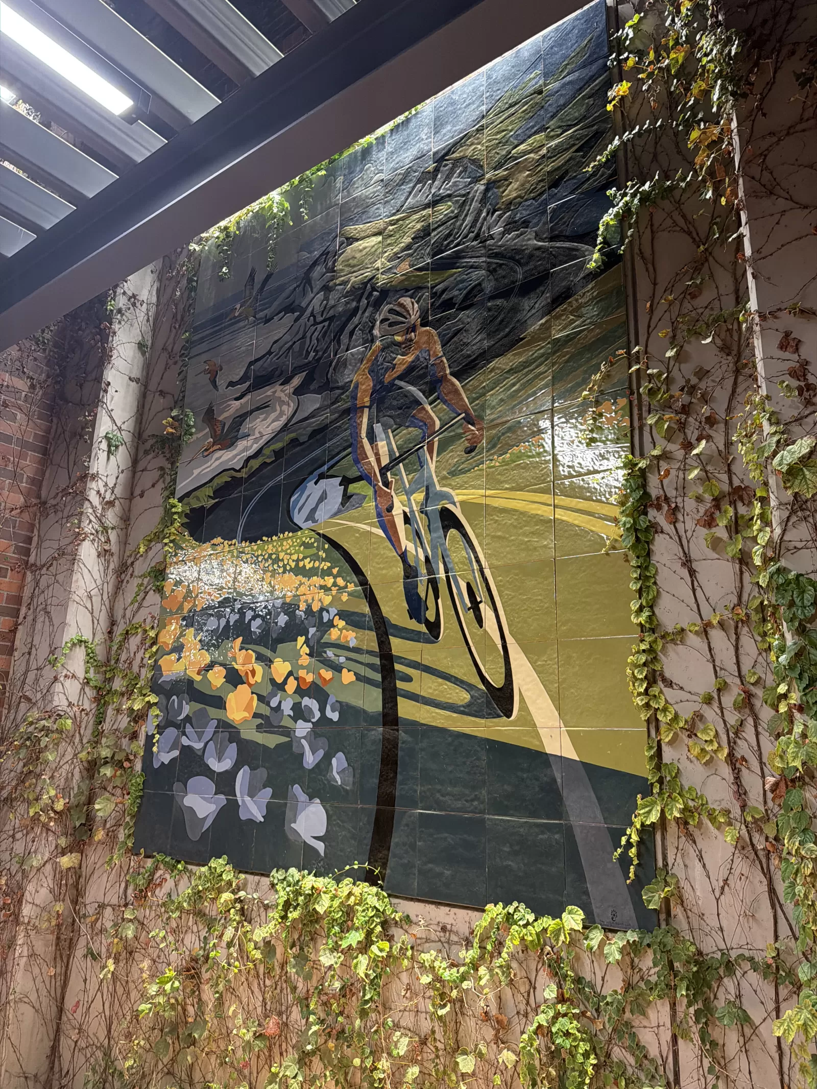 A large tiled mural depicts a helmeted cyclist riding through a stylized landscape with flowers, birds, trees, and sweeping green and blue forms. The mural is mounted on an exterior wall framed by climbing ivy and architectural beams.