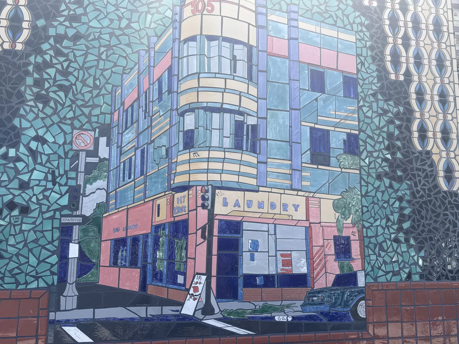 A stylized blue-and-pink city street scene centers on a curved storefront labeled “LAUNDRY,” with “COIN LAUNDRY” on a neighboring facade. The mural also shows buildings, streetlights, cars, plants, patterned geometric borders, and the number “305” near the top.