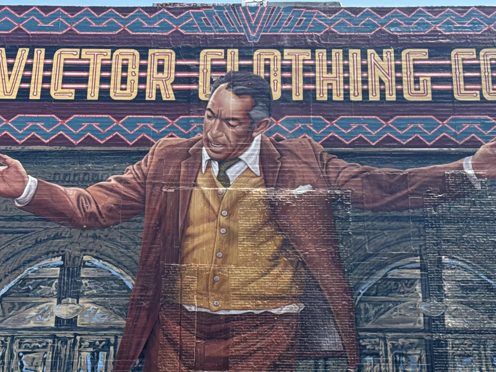A large wall mural shows a centrally positioned man in a brown suit, vest, and white shirt with his arms extended. Decorative geometric bands and partially visible large lettering reading “VICTOR CLOTHING” appear behind him, along with architectural details resembling a vintage storefront or building facade.