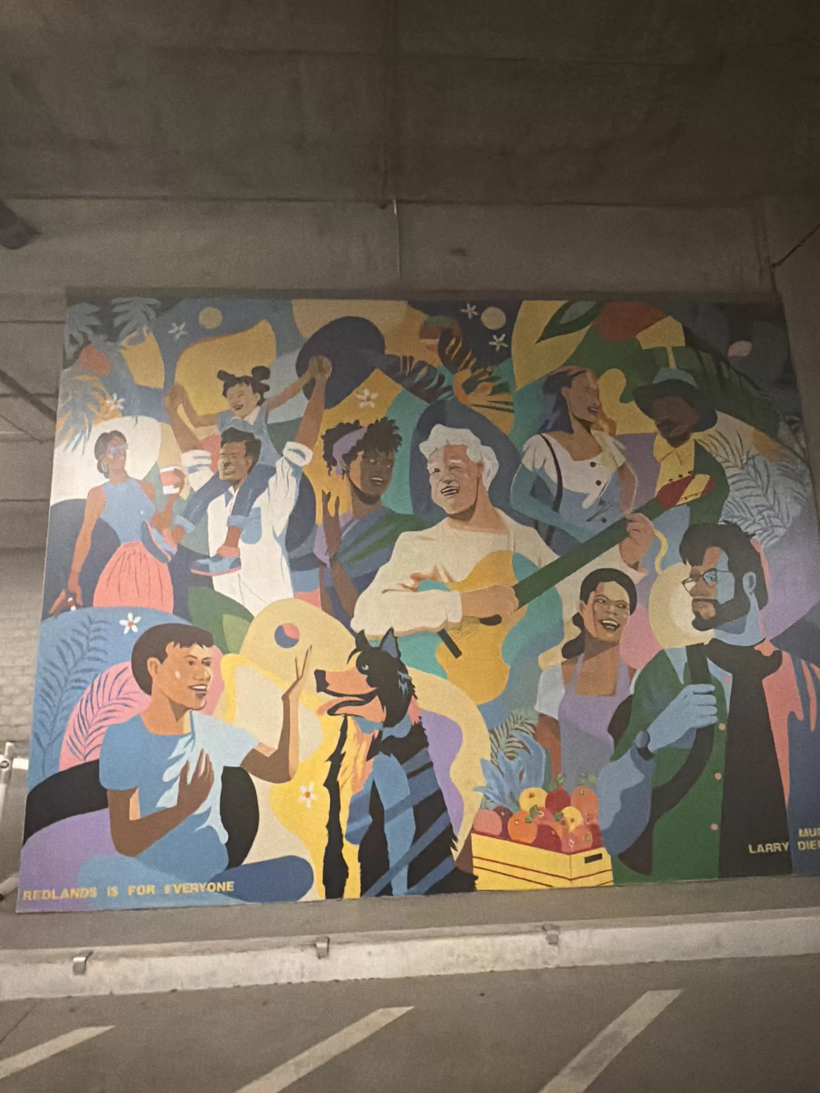 A colorful community mural portrays a diverse group of people gathered with musical instruments, food, plants, and a dog. The visible text along the bottom reads “REDLANDS IS FOR EVERYONE,” with partial lettering near the lower right edge.