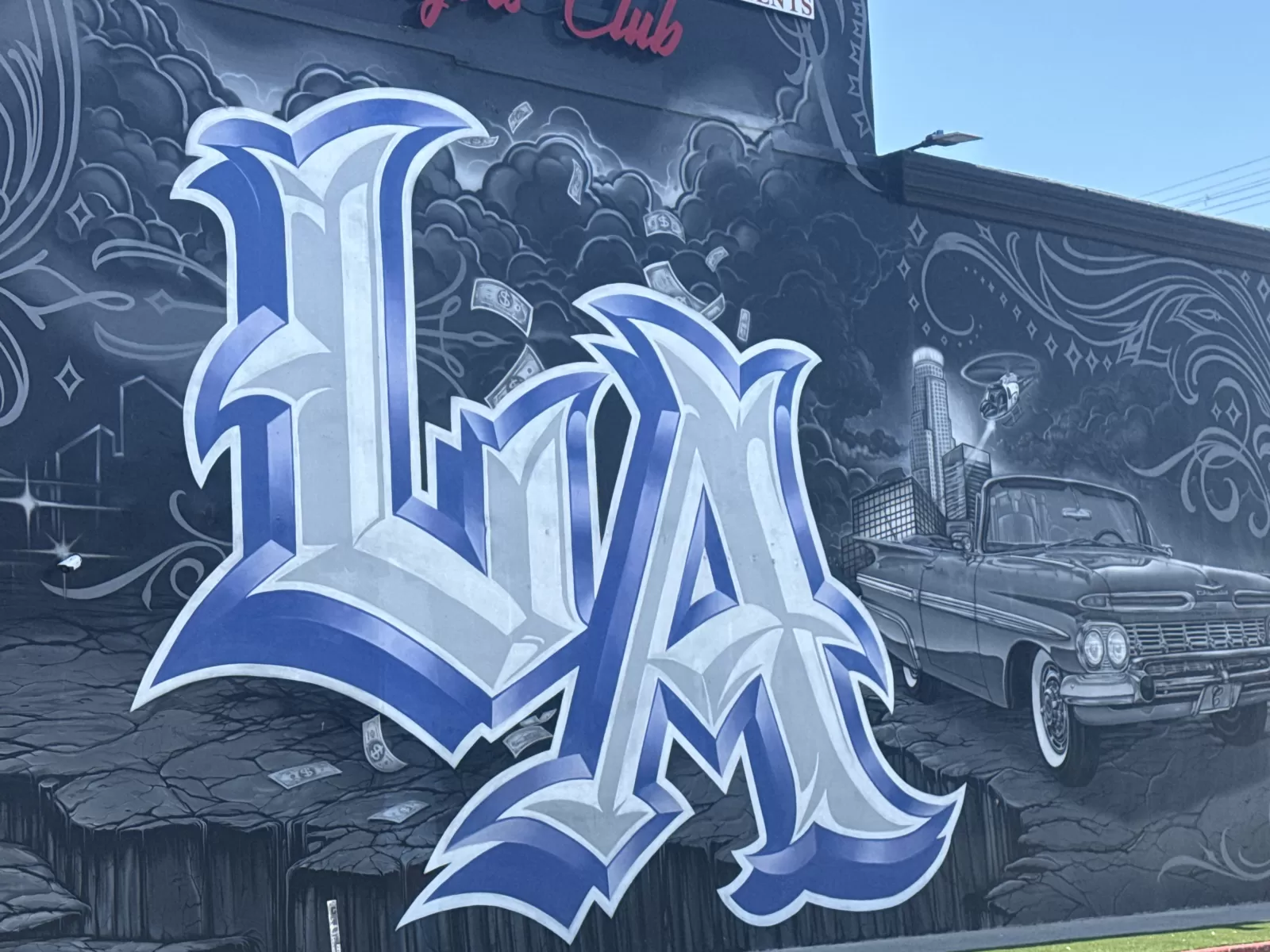 A large blue-and-white stylized graffiti-letter composition fills a dark wall, surrounded by cloud-like forms, decorative flourishes, and floating dollar bills. The mural also depicts a vintage car, a city skyline with a tall tower, and partial red lettering near the top that includes the word “Club.”