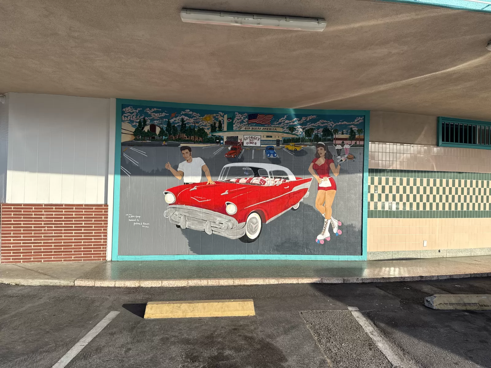 A painted storefront mural depicts a red vintage convertible, a carhop on roller skates, a man beside the car, and a retro diner parking lot scene. Visible text includes “GEORGE'S DINER” and “GOD BLESS AMERICA,” with an American flag above the diner.