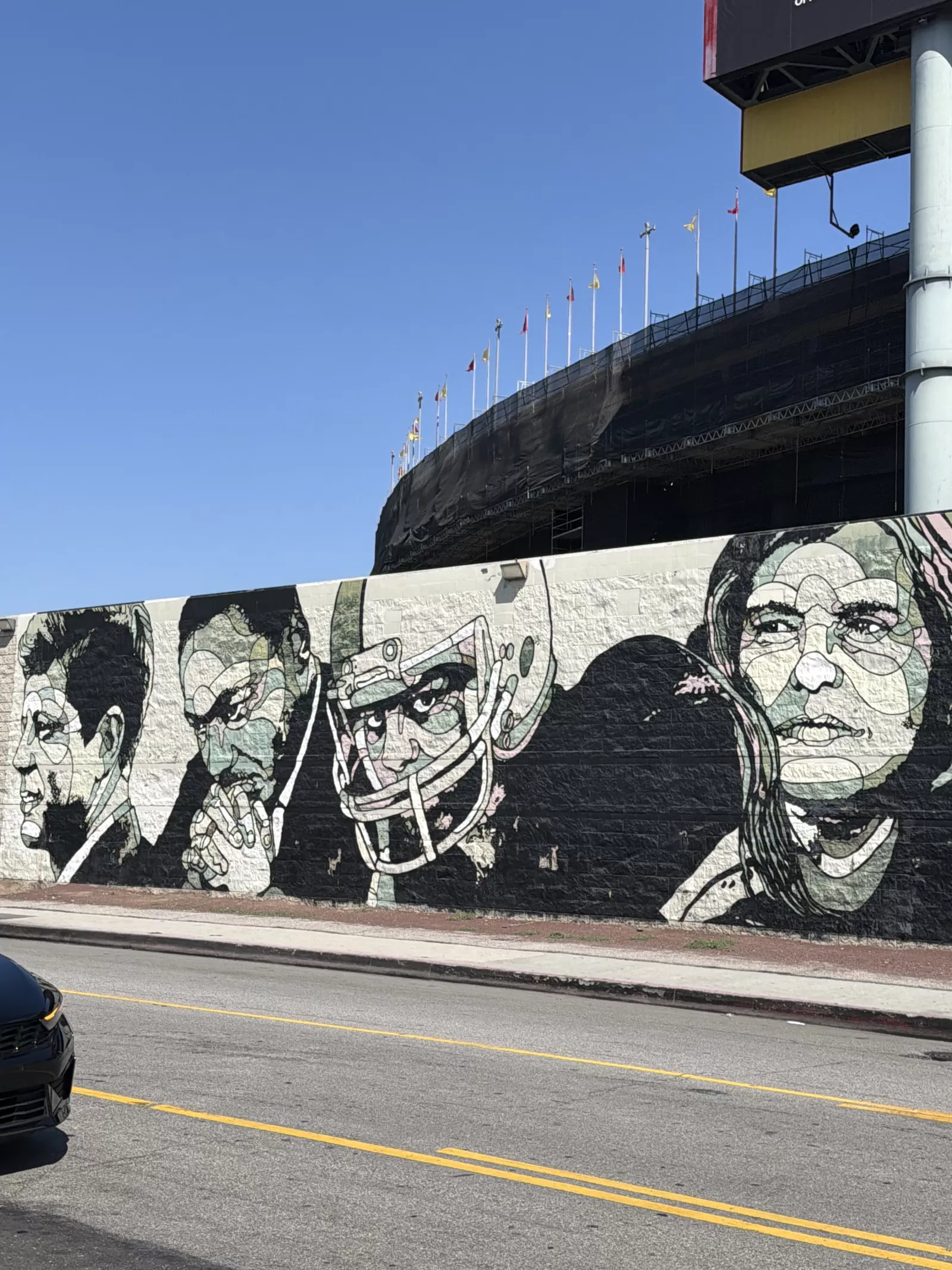 A large monochrome-and-muted-color mural covers a roadside wall with several stylized human portraits, including a central figure wearing a hat and a figure with long hair on the right. A large structure with flags rises behind the wall, and no clear mural text is visible.