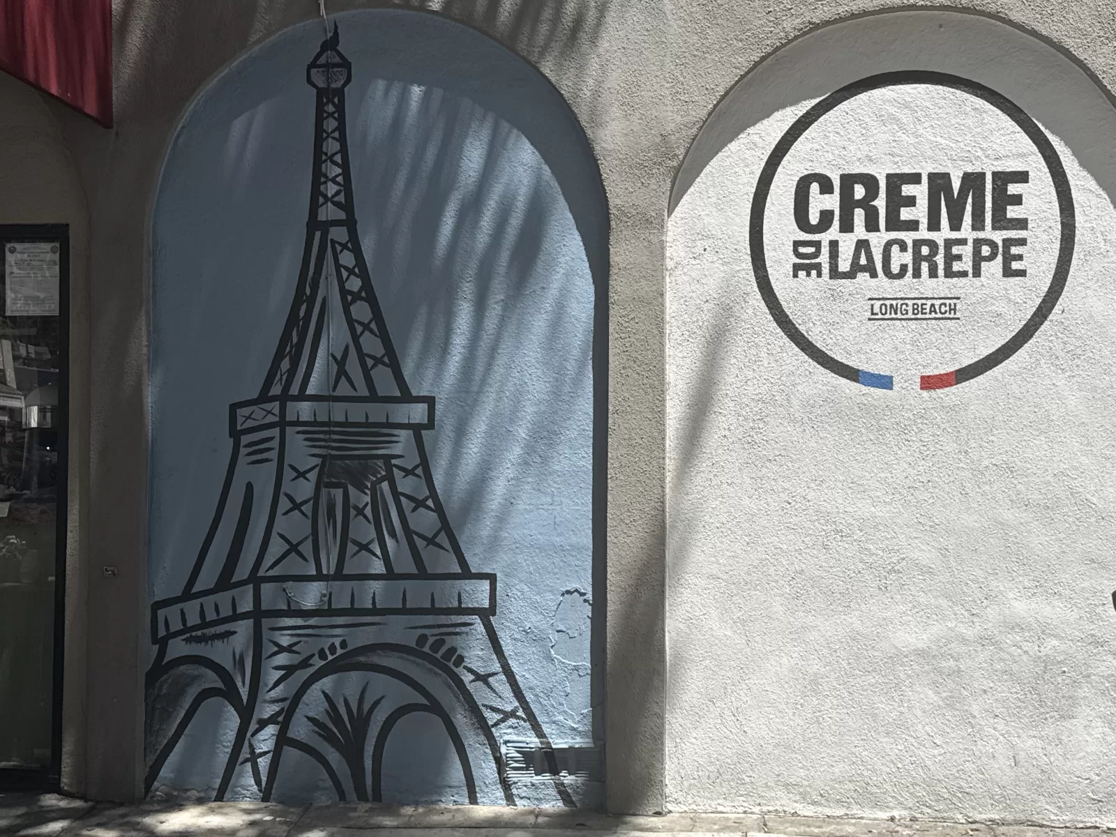 A black-and-blue painted Eiffel Tower fills an arched section of the storefront wall. Beside it, a circular sign reads “CREME DE LA CREPE” and “LONG BEACH.”