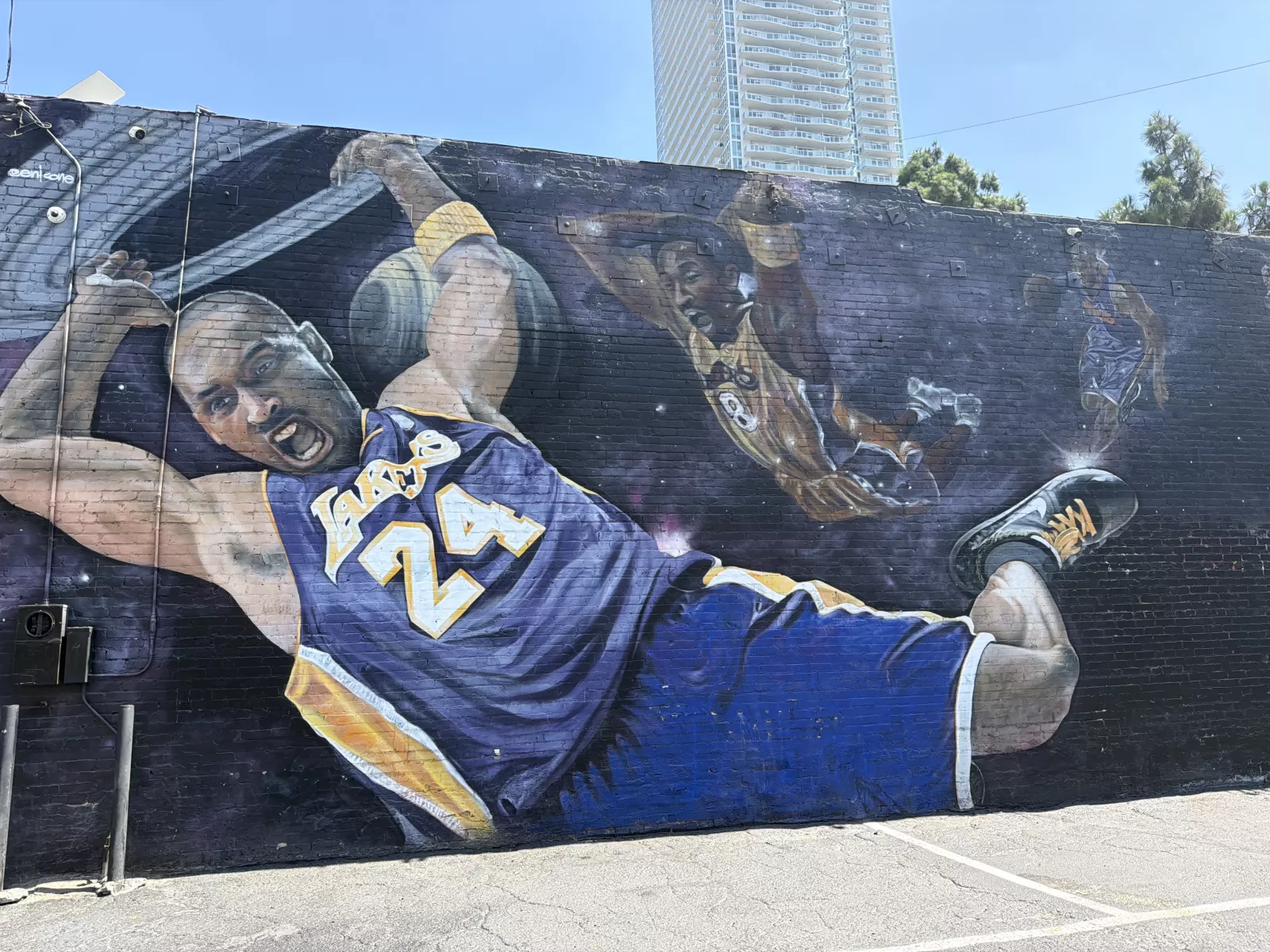 A large painted wall mural depicts multiple basketball players in action, including a foreground player wearing a purple-and-gold jersey with visible Lakers lettering and the number 24. The mural is painted on a dark brick wall beside a paved parking area, with a high-rise building and palm trees visible behind it.