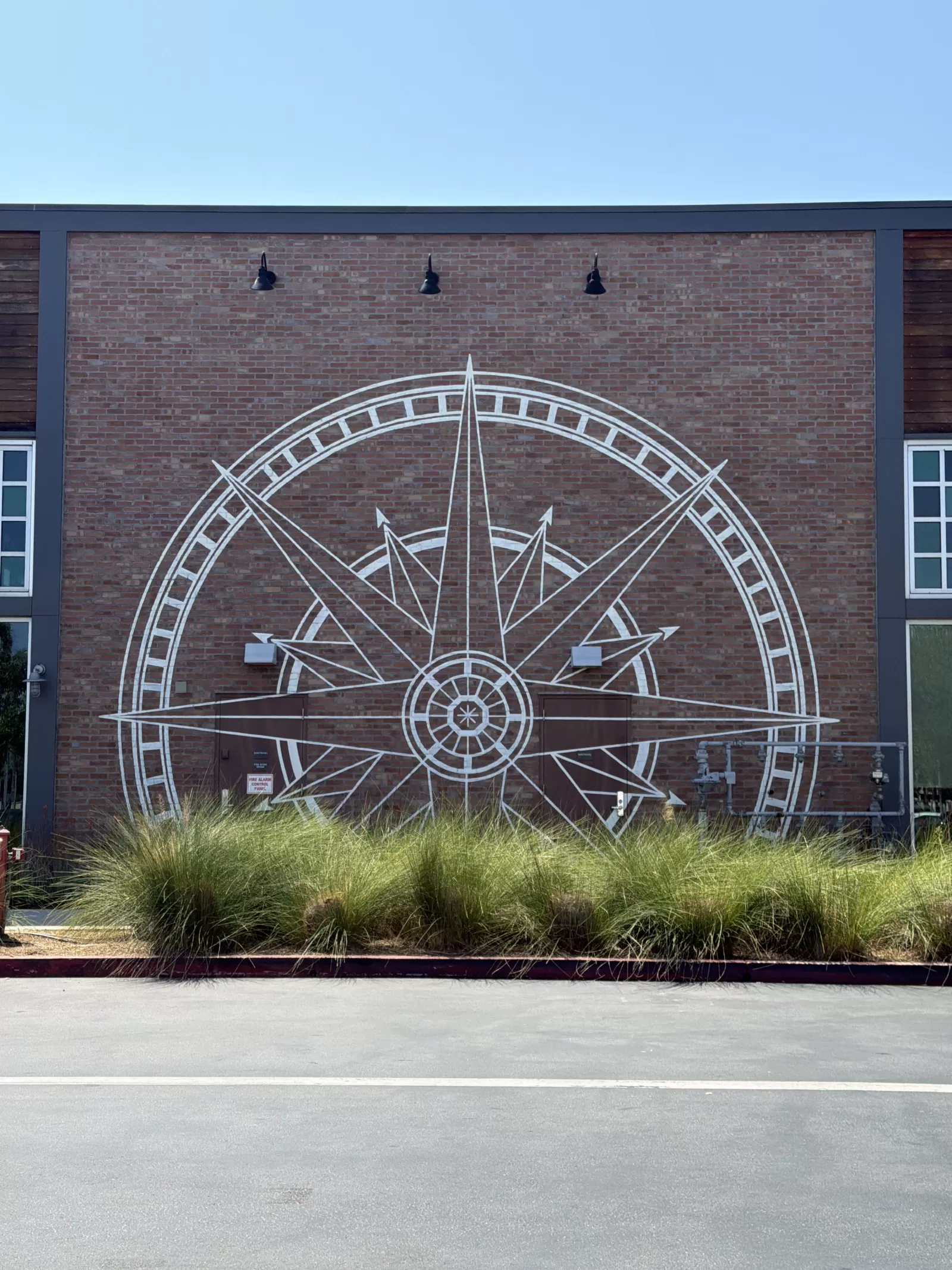 A large white compass-rose design with concentric arcs, directional points, and geometric details is painted on a red brick exterior wall. The mural is bordered by dark framing, windows, exterior lights, and landscaping.