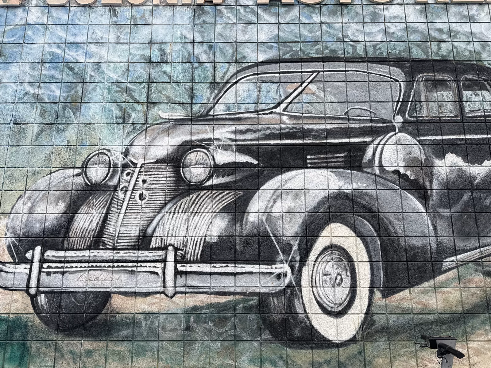 A large black-and-white mural depicts a vintage automobile with prominent headlights, grille, fenders, and wheels. The artwork is painted across a grid of square tiles with muted blue, gray, and green tones in the background.